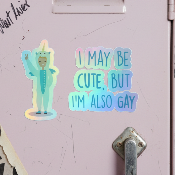 Cute But Gay Holographic Stickers on a pastel pink locker: one with a waving unicorn onesie queer cutie, one reading “I may be cute, but I’m also gay.” Silver latch and paper note edge in view.