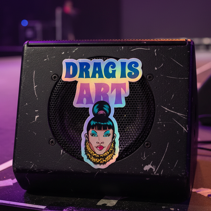 Black speaker with visible scrapes on a dim stage. A holographic "Drag Is Art" sticker shows bold drag makeup, earrings, and a high bun.