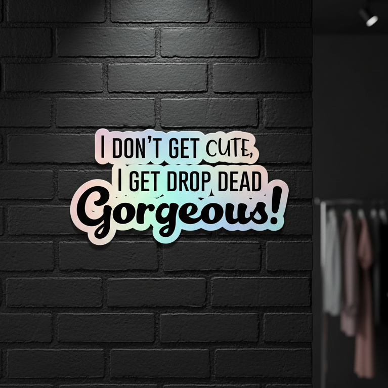 Drop Dead Gorgeous holographic stickers shimmer by a pastel sign on a dark brick wall reading, “I don’t get cute, I get drop dead gorgeous!” A clothes rack sits in soft shadows.