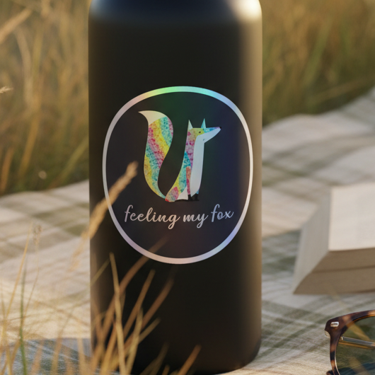 Holographic Feeling My Fox stickers with queer vibes, beside a black bottle with a colorful fox and "feeling my fox" text, on a checkered blanket in tall grass, sunglasses and book nearby in soft daylight.