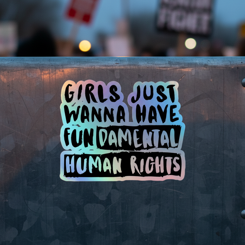 Holographic "Girls Just Wanna Have Fundamental Human Rights" sticker with bold, vibrant colors on metal, rally signs blurred behind.