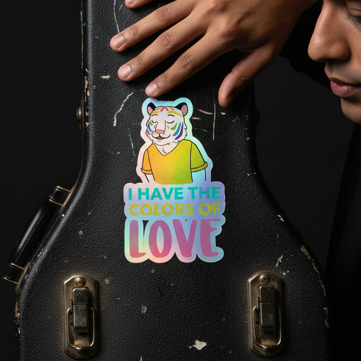 Holographic sticker on a black guitar case shows a rainbow tiger in a yellow tee; text reads, I HAVE THE COLORS OF LOVE. Hand visible.