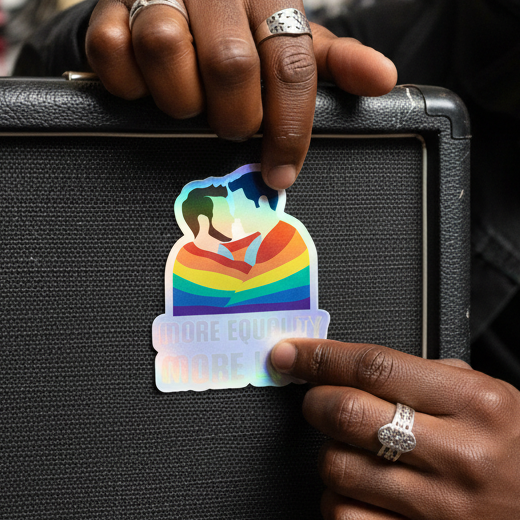 Holographic sticker with rainbow colors, two people kissing, and “MORE EQUITY” in blue and white, placed by a dark-skinned hand with silver rings on a black surface.