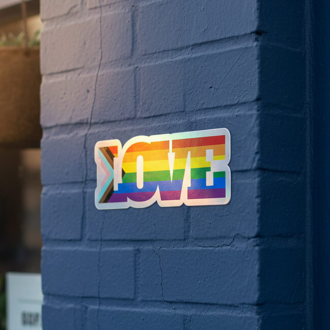 Progress LGBT holographic vinyl sticker with "LOVE" in rainbow flag colors and a > symbol, stuck on a blue brick wall.