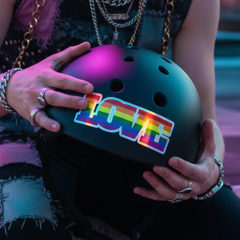 Black helmet featuring a "Gay Love" holographic sticker, held by someone in ripped jeans and rings, under bold pink-blue lighting.