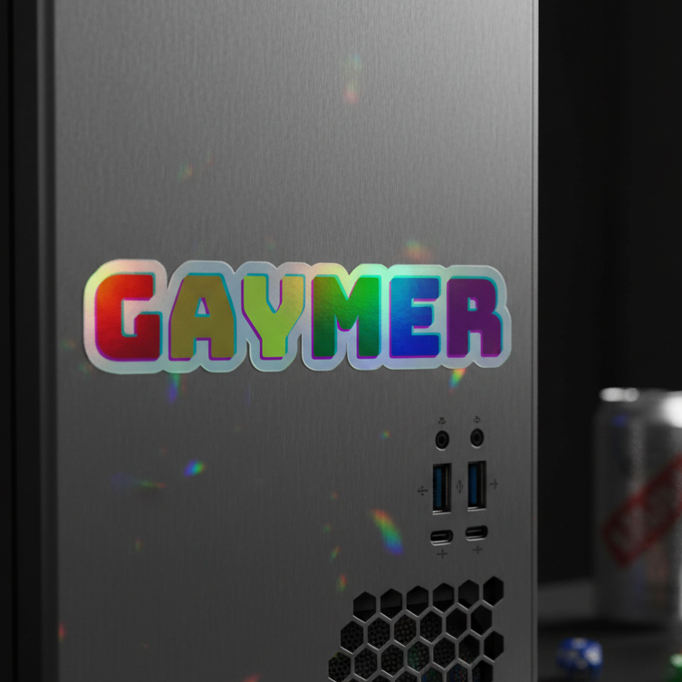 Metallic PC tower rocking a shiny holographic "Gaymer" sticker, bold letters. Two USB ports, two audio jacks, vent underneath. Blurred can and blue die in back.