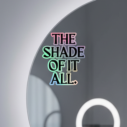 Round mirror with bold, colorful “THE SHADE OF IT ALL.” text; blurred neutral background and a round light reflected at lower right.