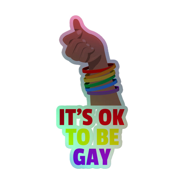 A dark-skinned hand forms a finger-heart gesture, adorned with rainbow bracelets, above bold text: IT'S OK TO BE GAY. It's Ok to Be Gay Holographic Stickers celebrate queer pride and self-expression.