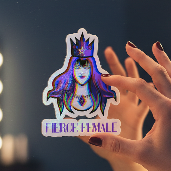 Fierce Female Bubble-Free Sticker showing a crowned woman with vibrant hair, bold makeup, and “FIERCE FEMALE” below. A hand with dark red nails holds it against a blurred mirrored or glass background.