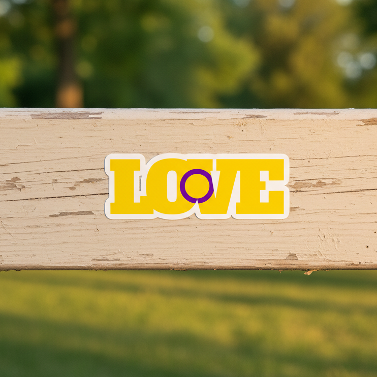 Bubble-free sticker says “LOVE” in bold yellow vinyl; the “O” is a purple intersex ring. It sits on light wood, greenery aglow.