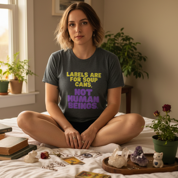 Labels Are For Soup Cans tee on a queer femme sitting cross-legged on a bed amid crystals, books, and cards