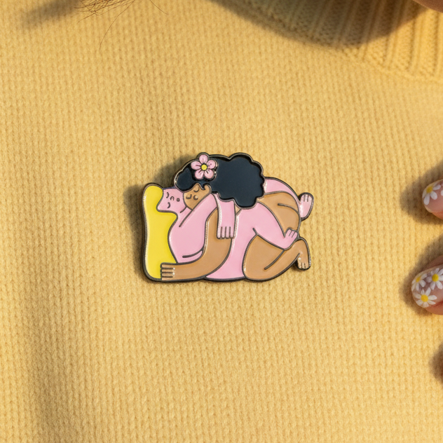 Close-up of a flower enamel pin showing two intertwined figures in pink and yellow