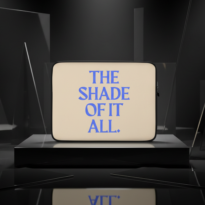 Beige "The Shade Of It All" laptop sleeve with bold blue text, standing on a black platform among sharp shapes in moody lighting.