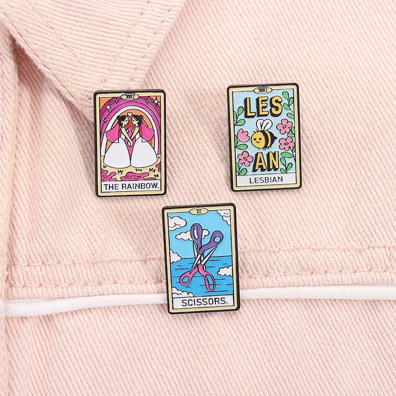 Three lesbian tarot enamel pins reading The Rainbow, Lesbian, and Scissors on pink fabric