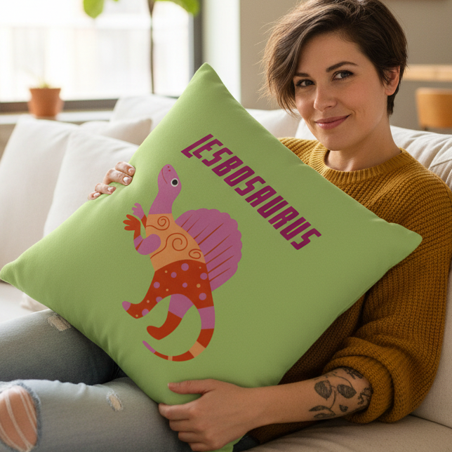 A person in a mustard sweater holds the Lesbosaurus Pillow—colorful cartoon dinosaur, bold "LESBOSAURUS" text