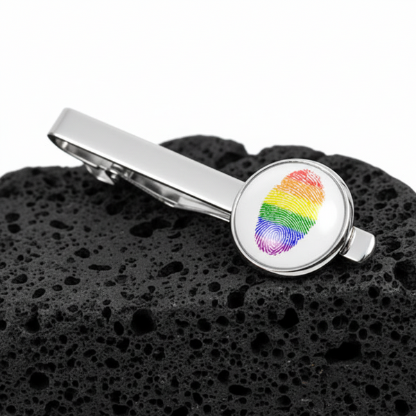 Silver LGBT tie clip with a rainbow fingerprint at its round end pops against textured black volcanic rock—a proud