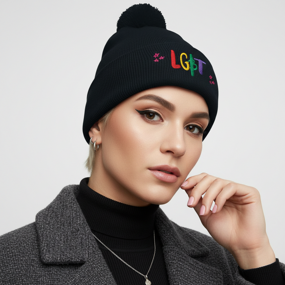 LGBT Pom Pom Beanie, black knit with “LGBT” in rainbow thread, worn by a person in a gray coat, hoop earrings, winged liner
