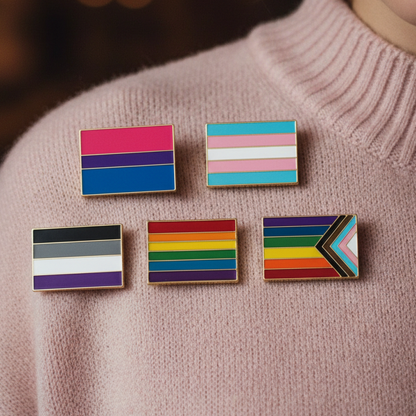 Five enamel pride flag pins arranged on a pale pink ribbed sweater