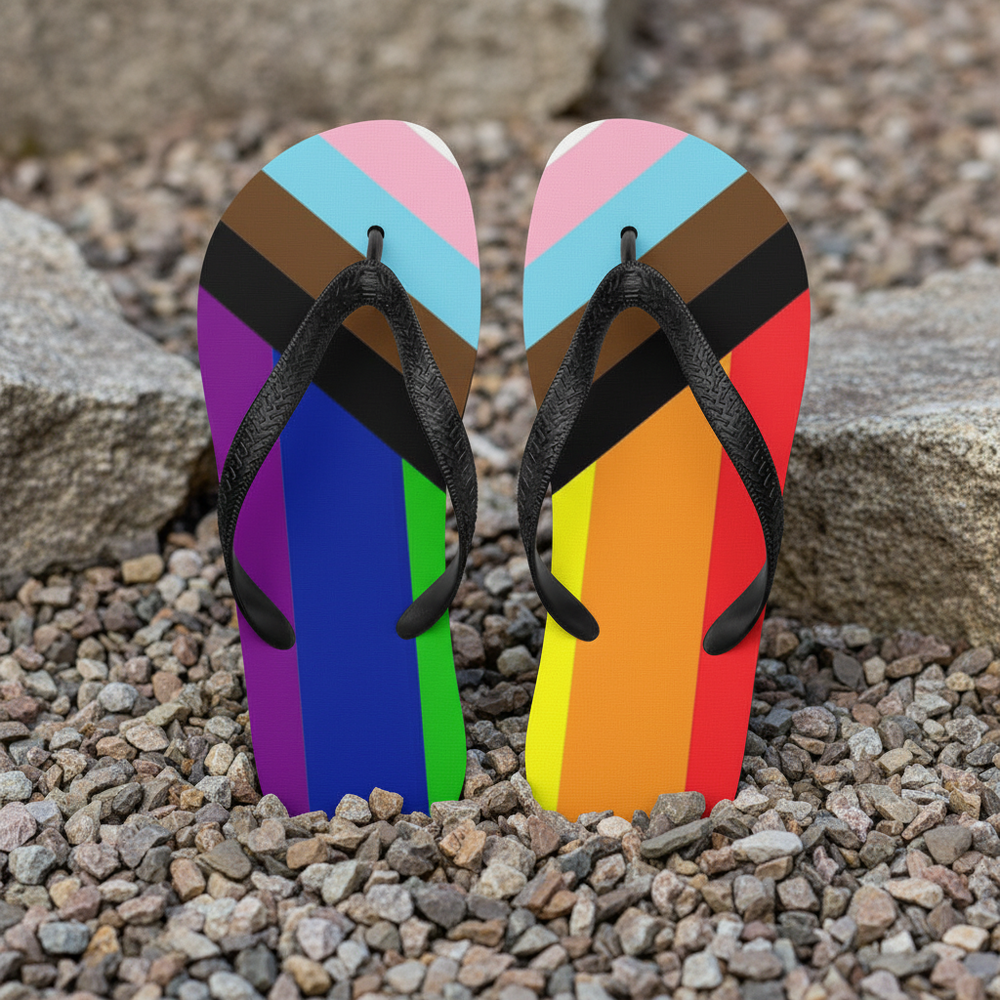 Progress Pride flag flip-flops with black straps rest on small rocks, footbeds bursting with queer colors