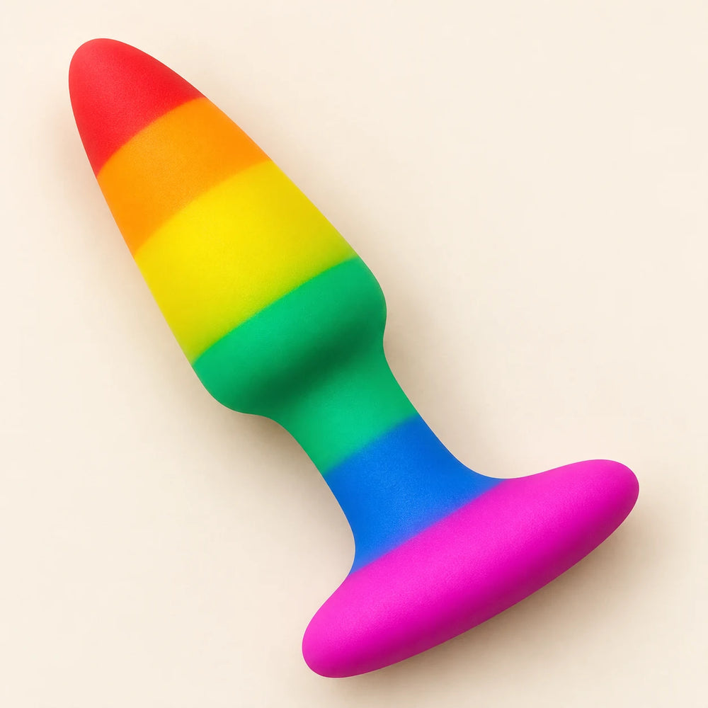 Rainbow-striped silicone butt plug lying diagonally on ivory, showing its tapered shape and wide pink flared base.