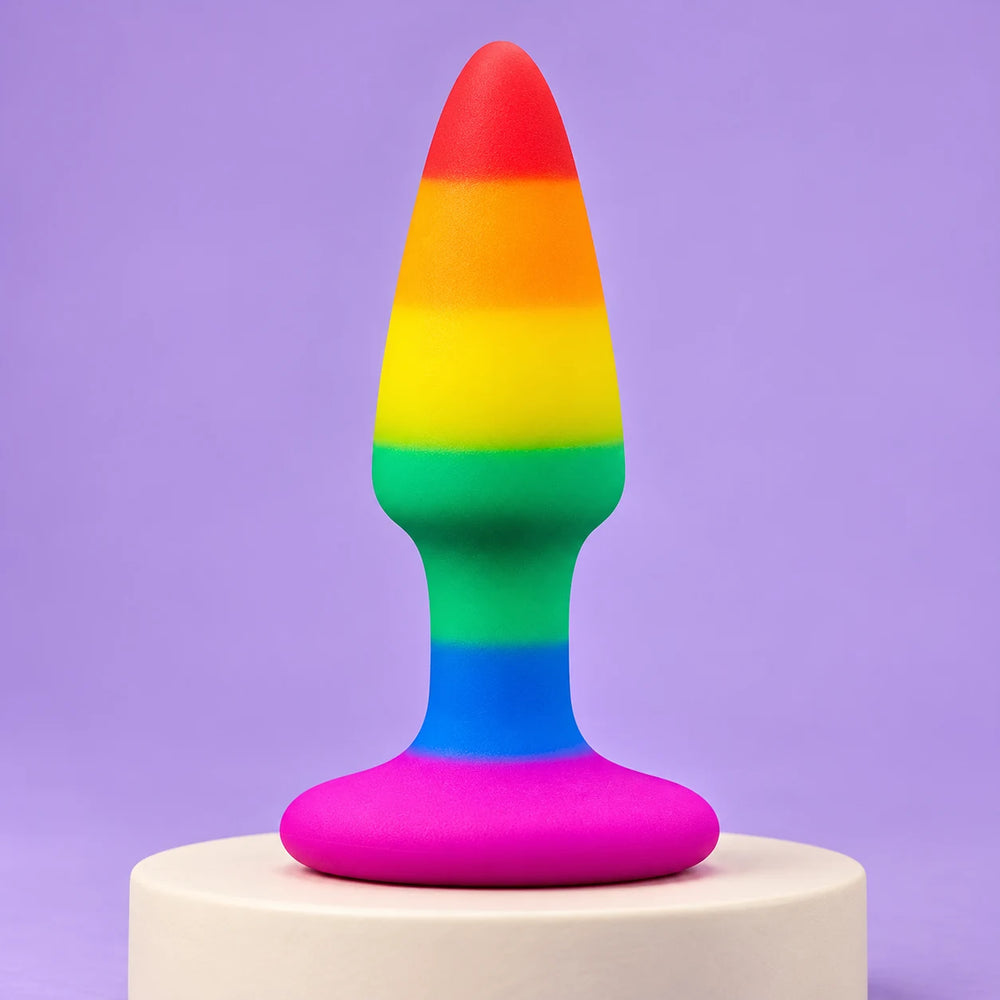 Rainbow silicone butt plug with a red tip, blue neck and wide pink flared base standing on an ivory plinth against lavender.