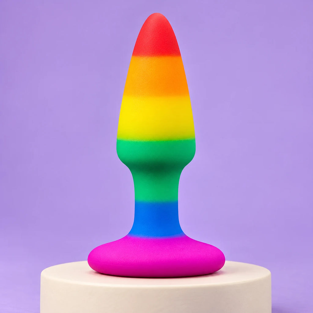 Rainbow silicone butt plug with a red tip, blue neck and wide pink flared base standing on an ivory plinth against lavender.