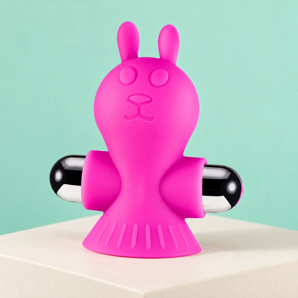 One pink rabbit-shaped Liberating Love vibrating nipple clamp with rounded black ends on an ivory block against mint.