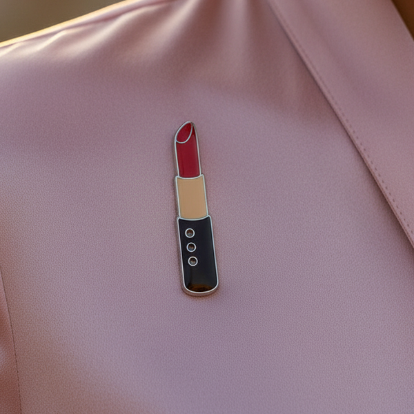 Lipstick-shaped enamel pin with red, nude, and black sections on a pink surface