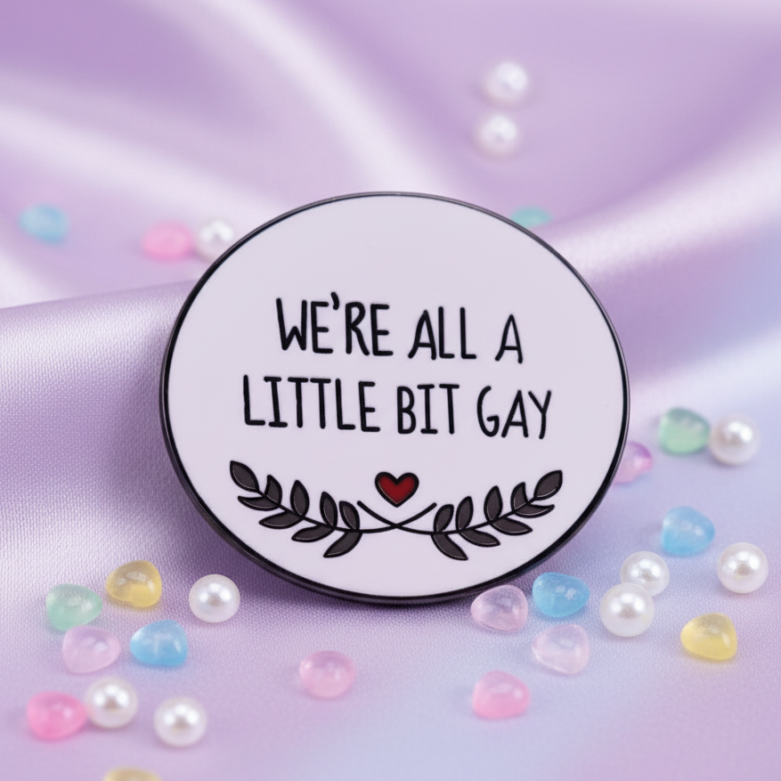 Close-up enamel pin reading “WE’RE ALL A LITTLE BIT GAY,” with a red heart and leafy border