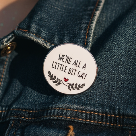 Enamel pin reading “WE'RE ALL A LITTLE BIT GAY” on a denim jacket