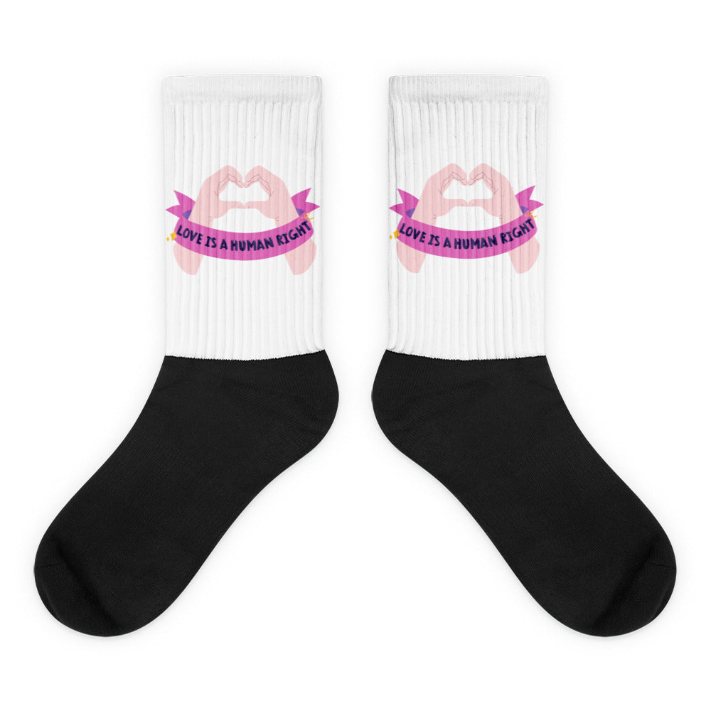 Pair of black-and-white crew socks with pink hand-heart graphics and “LOVE IS A HUMAN RIGHT” text