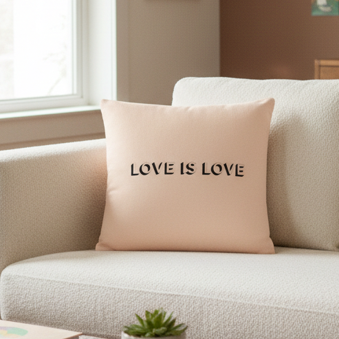 Light beige "LOVE IS LOVE" pillow with bold black text on cream sofa, beside a green succulent, in soft window light