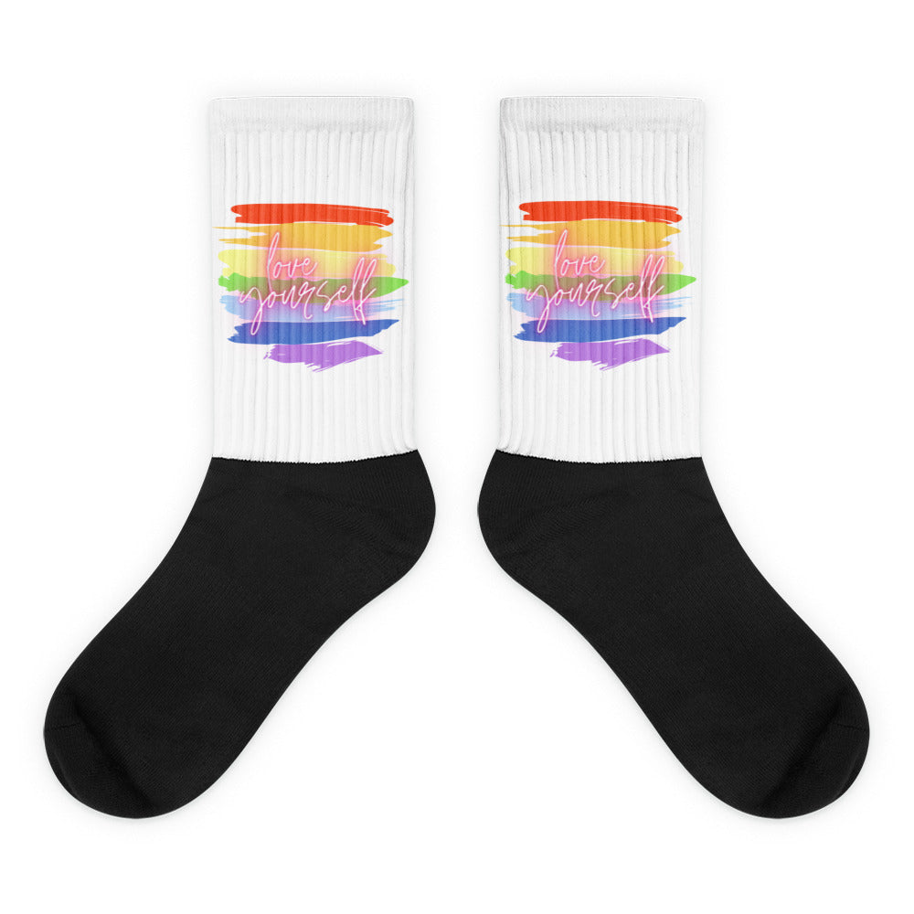 Pair of crew socks with black feet and white ribbed cuffs bearing rainbow stripes and “love yourself”