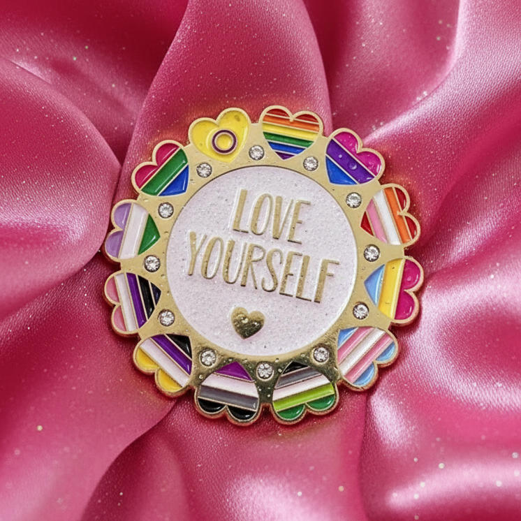 Close-up of a rainbow heart enamel pin reading “LOVE YOURSELF” on pink satin