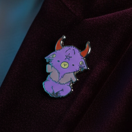 Purple goat enamel pin with curved horns and yellow eyes on dark fabric