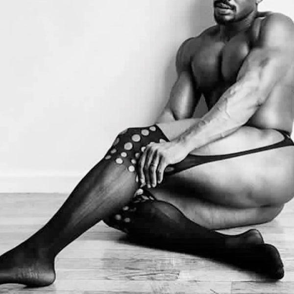 Black-and-white photo of a seated shirtless person wearing sheer thigh-high stockings with large dots