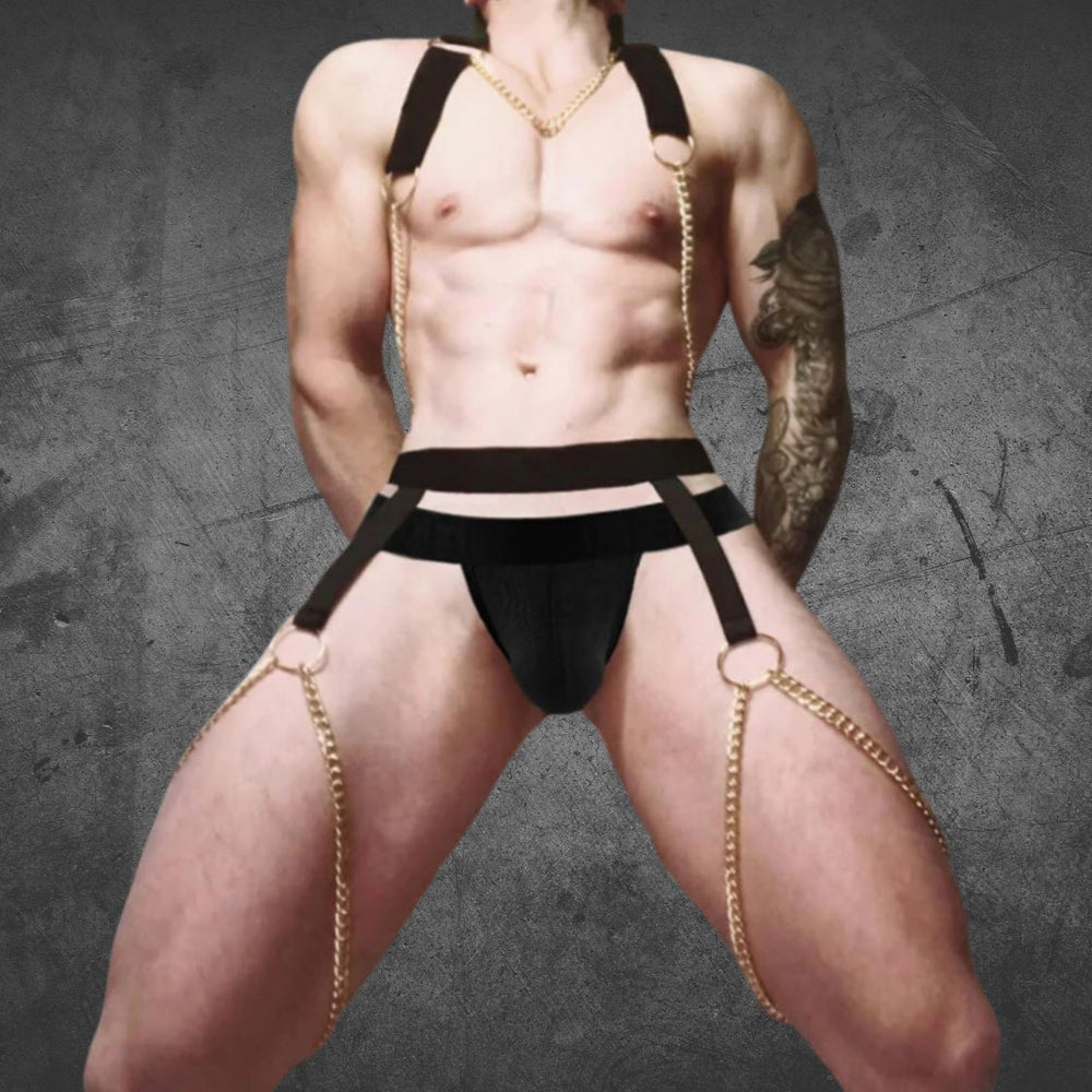 Front view of a shirtless model wearing a black halter harness with gold chains and a matching jockstrap