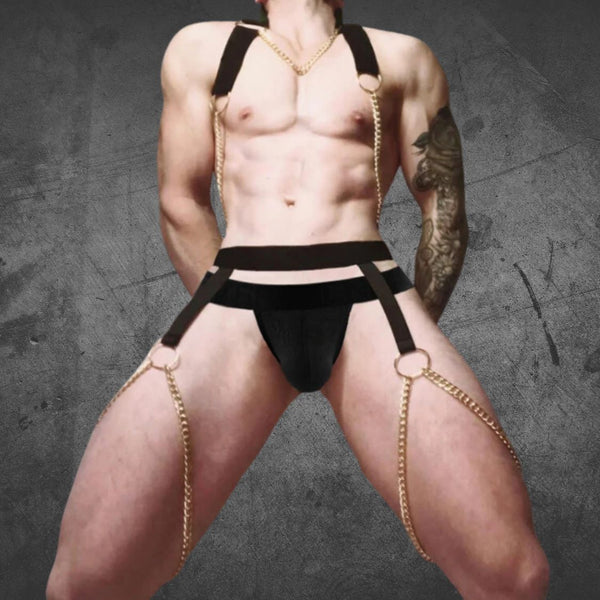 Front view of a shirtless model wearing a black halter harness with gold chains and a matching jockstrap