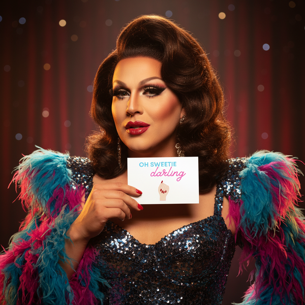 Drag performer holding an “OH SWEETIE darling” postcard in a sequined, feather-trimmed costume