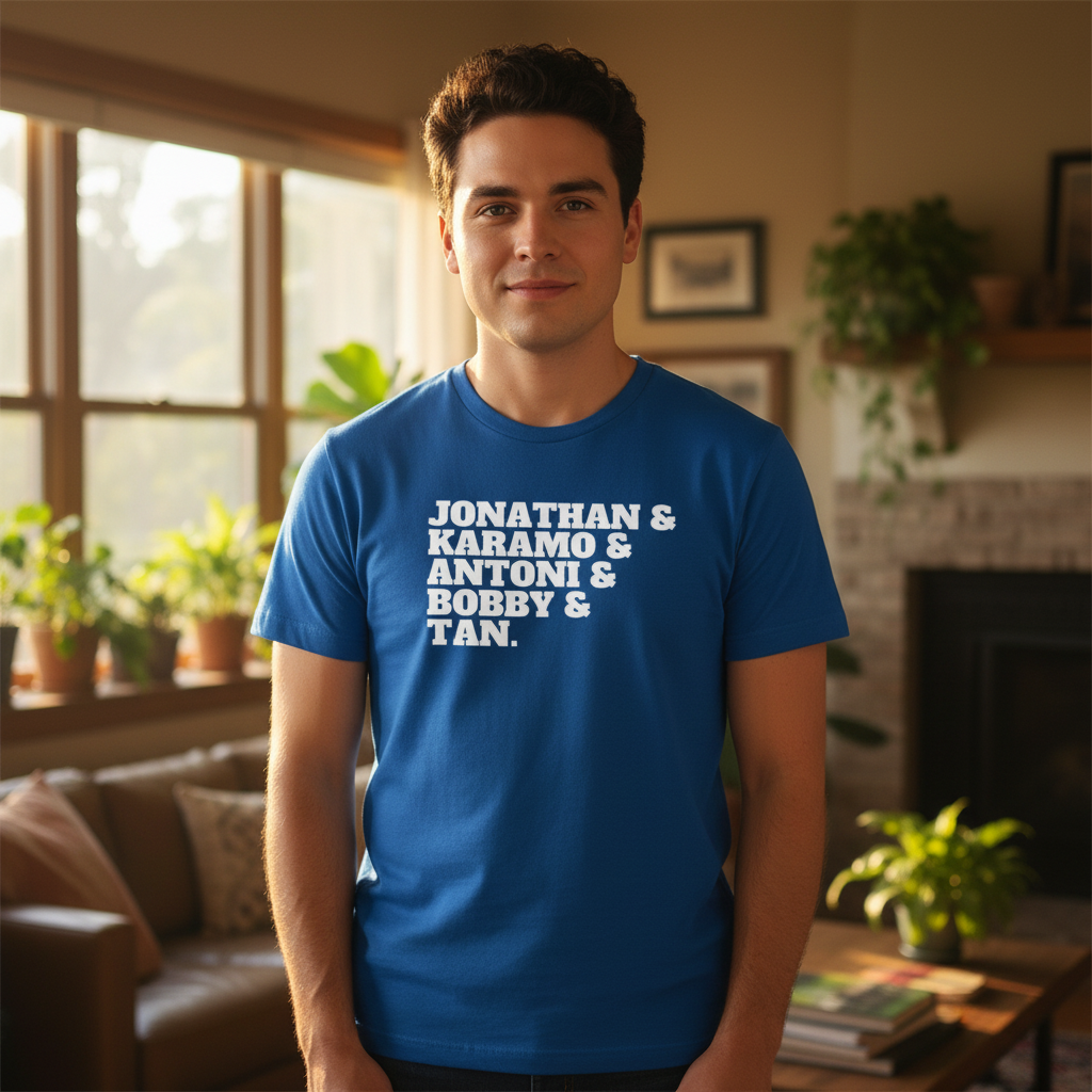 Blue T-shirt with bold white "Jonathan & Karamo & Antoni & Bobby & Tan" text, worn by a smiling young queer guy indoors.