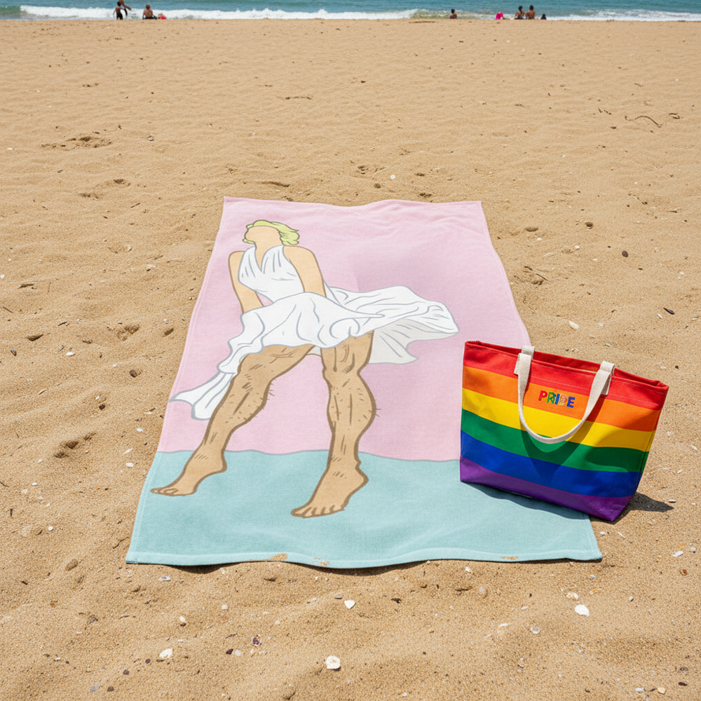 The Daddy Monroe Towel with a white-dressed figure in Monroe’s pose next to a rainbow-striped "PRIDE" tote at the beach.