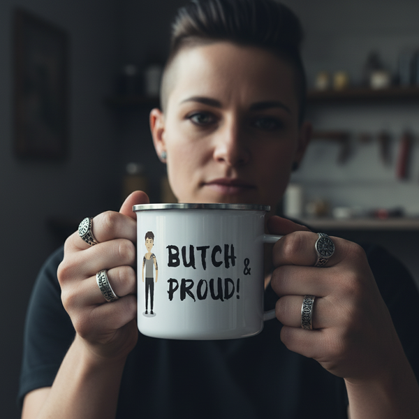 Person with an undercut holds a Butch & Proud enamel mug featuring a cartoon figure. Their silver rings add flair, embodying pride and self-expression, in line with Queer In The World's inclusive spirit.