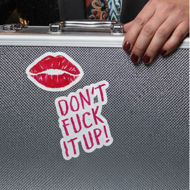 Silver textured briefcase with "Don't Fuck It Up!" queer stickers—bold red lips, pink slogan—held by maroon-manicured hand.