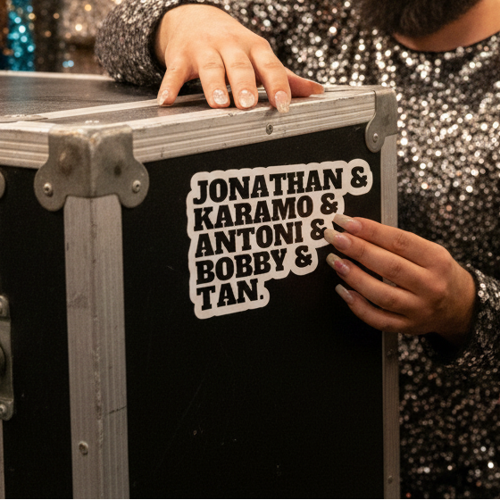 Black case decked out with "Jonathan & Karamo & Antoni & Bobby & Tan" stickers, held by a bearded person in sequins.