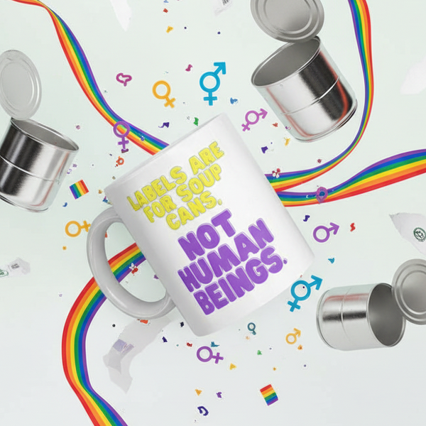 White mug reading “Labels Are for Soup Cans, Not Human Beings” among rainbow ribbons and symbols