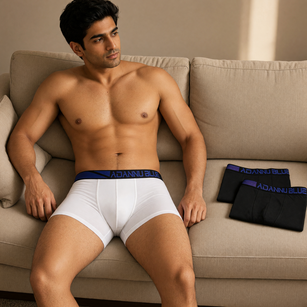 Model seated on a beige sofa in white ADANNU BLUE boxer briefs, with two folded black pairs beside him
