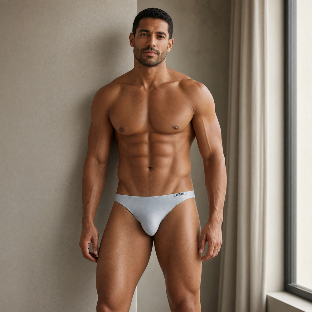 Model standing beside a window in shimmering pale silver briefs with a small TAUWELL side label