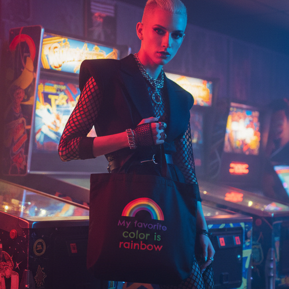 Person holds a black tote reading “My favorite color is rainbow” in a neon-lit arcade