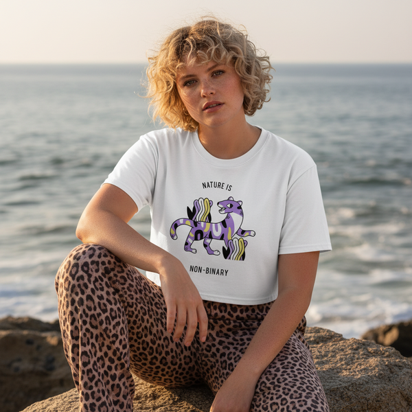 Model seated by the sea wearing a white gay crop top with a purple creature and “NATURE IS NON-BINARY”