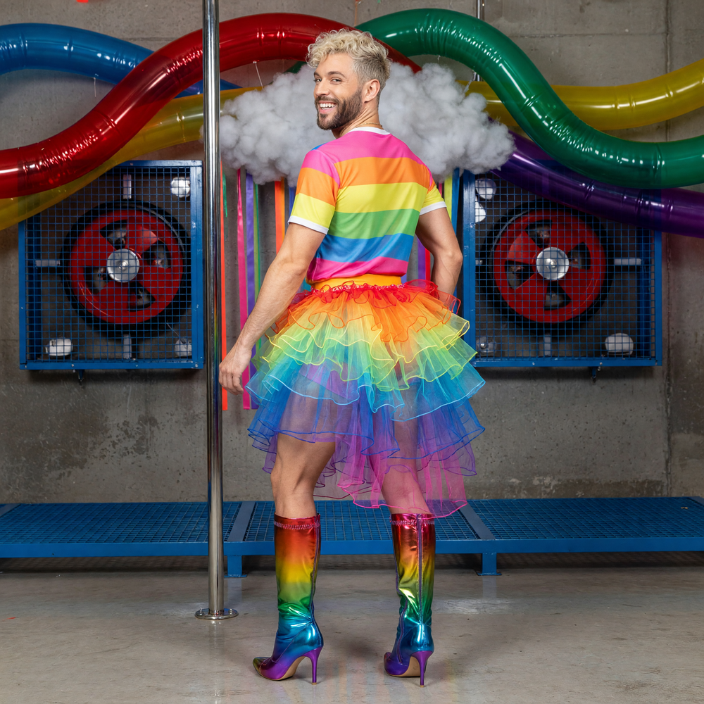 Rear three-quarter view of a model wearing the Neon Rainbow Tutu with cascading rainbow tulle ruffles concentrated at the back, in a colourful industrial set.